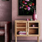 A framed floral art print with vibrant pink, red, and purple flowers on a black background, displayed on a dusty pink wall above a wooden sideboard in a styled room.
