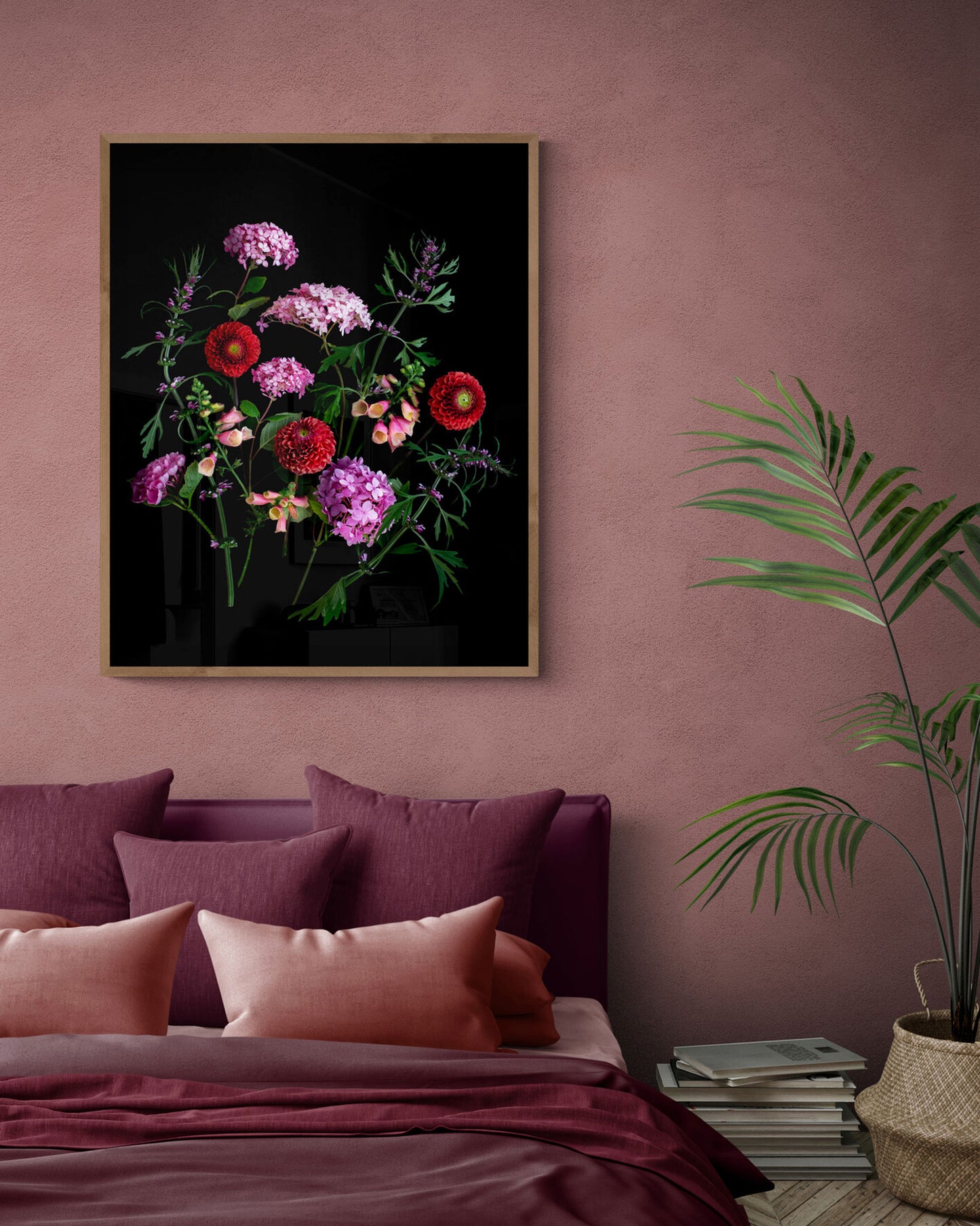 A framed floral wall art print hangs above a bed in a styled bedroom. The artwork shows a bouquet of pink, red, and purple flowers on a black background, with a gold-toned frame.