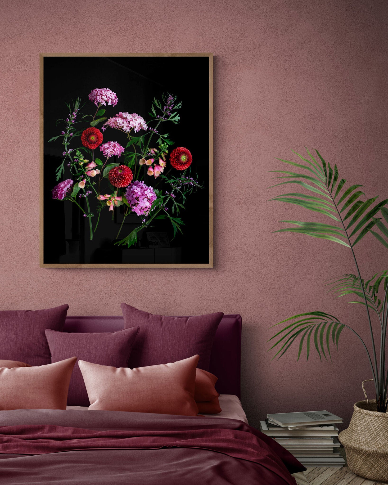 A framed floral wall art print hangs above a bed in a styled bedroom. The artwork shows a bouquet of pink, red, and purple flowers on a black background, with a gold-toned frame.