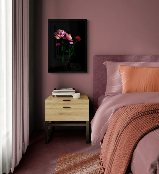 A framed wall art print featuring peach pink double tulips with Ranunculus  on a black background, styled in a bedroom setting above a wooden nightstand next to a bed with dusty rose bedding and a rust-colored throw blanket.